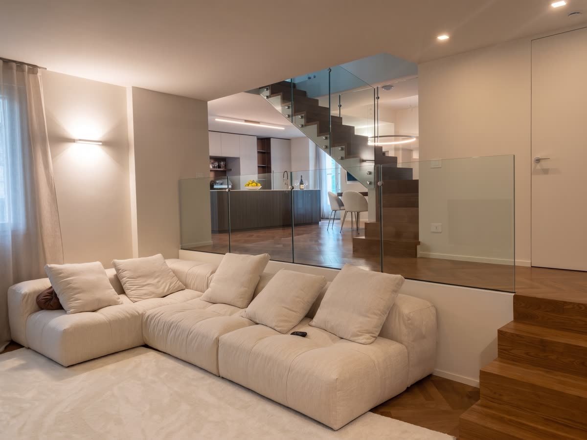 Soggiorno con divano componibile chiaro e scala con parapetto in vetro